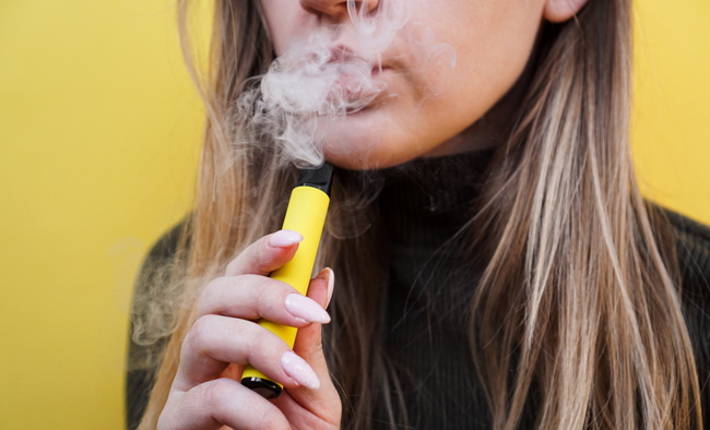 A woman holds a disposable vape while exhaling vapour.